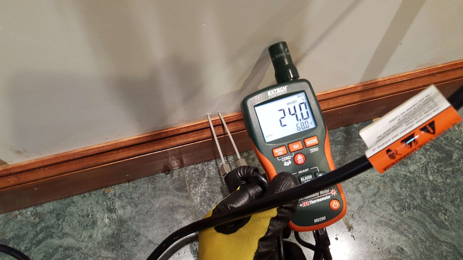 Extech pin-type moisture meter showing 24 percent reading at baseboard indicating wet wall